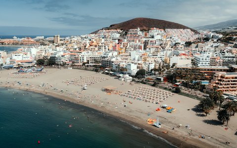 Aerial view of the sandy beach and white town of Los Cristianos below a brown hill, beside a blue sea