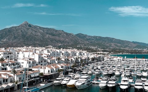 Aerial view of a marina full of white yachts in front of white buildings, with a mountain behind under a blue sky