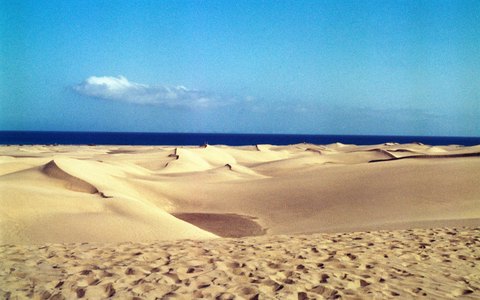 Golden sand dunes rolling towards a distant blue sea under a clear blue sky with a few clouds