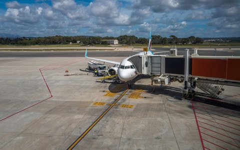 Verkehrsflugzeug an einer Fluggastbrücke auf dem Vorfeld des Flughafens Palma, dahinter Bäume und Wolken