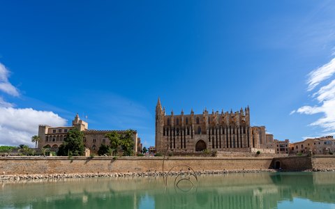 Palma Cathedral and the Almudaina Palace above the seafront walls, reflected in still water under a blue sky
