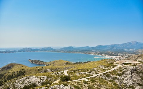 A wide view over Alcudia Bay with green headlands, the coastline and mountains under a clear blue sky