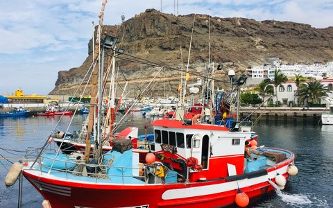 A red and white fishing boat moored in Mogan harbour below a rocky hill, under a blue sky