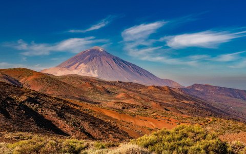 The peak of Mount Teide rising above red and brown volcanic ground, under a deep blue sky with white clouds
