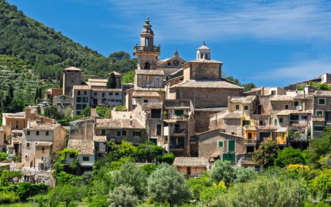 The stone village of Valldemossa and its church tower among green trees, under a blue sky