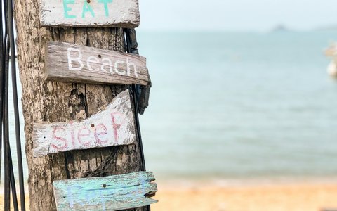 Hand-painted wooden signs reading Eat, Beach and Sleep on a tree trunk, with sand and calm sea behind on Bophut Beach
