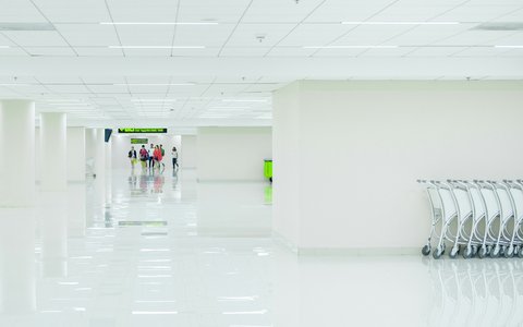 A long, bright white terminal corridor at Don Mueang Airport, with luggage trolleys lined up on the right and a few travellers walking towards a sign