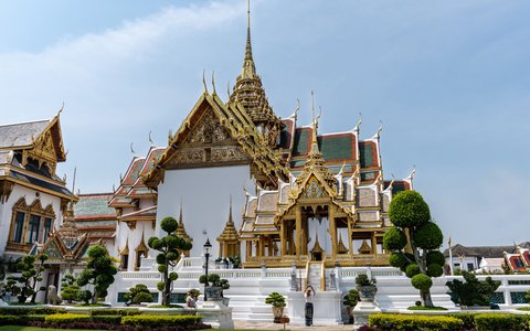 Gilded spires and white halls of the Grand Palace in Bangkok behind clipped trees and lawn