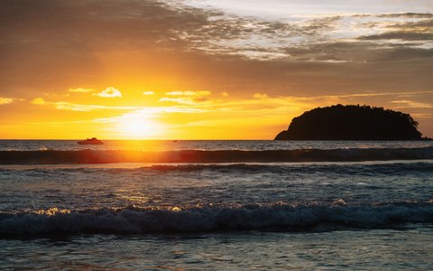 The sun setting behind a small island off Kata Beach, with waves rolling in under a golden sky