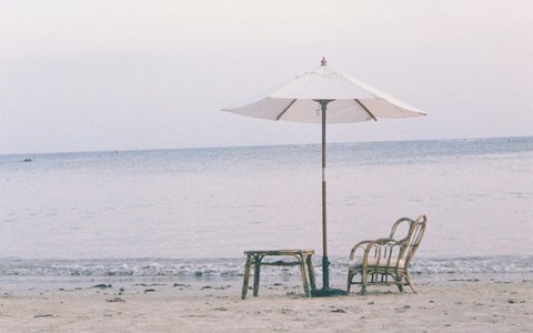 A white parasol over a wooden chair and a small table on the sand at Lamai Beach, with calm sea and a pale sky behind