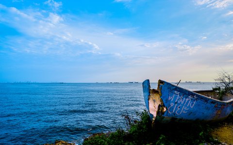 An old blue boat beached on the Pattaya shore with the blue sea and a bright sky with white clouds
