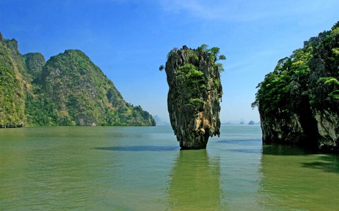 A narrow limestone pillar rising from green water between steep forested karsts in Phang Nga Bay, under a blue sky