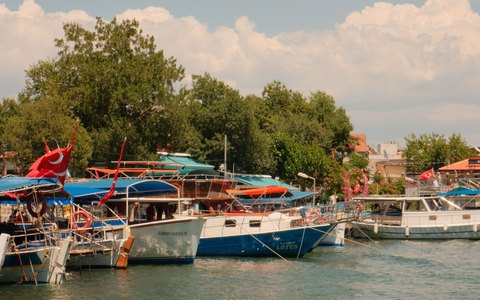 Painted tour boats flying Turkish flags moored along a green river bank at Dalaman, under a sky with white clouds