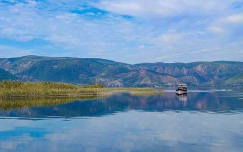A calm lake reflecting reed beds and wooded hills near Dalyan, under a blue sky with white clouds