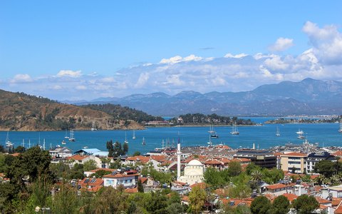 Aerial view of Fethiye town, harbour and bay with mountains behind, under a blue sky with white clouds