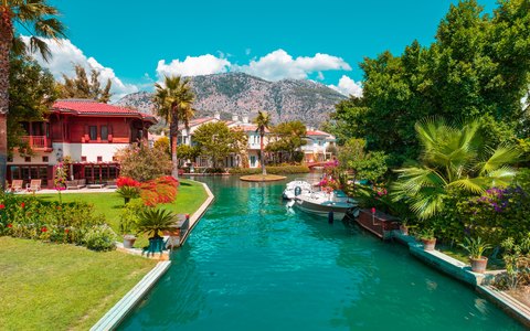 A turquoise canal lined with palm trees, villas and moored boats at Gocek, with mountains behind