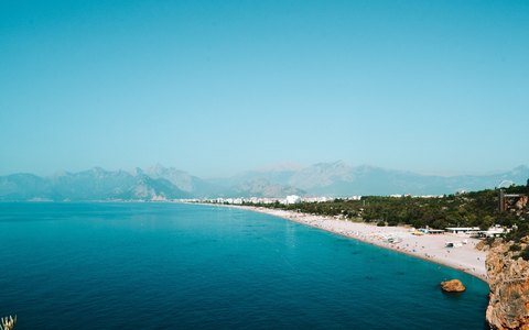 Aerial view of the long pebble beach of Konyaalti curving along turquoise water, with mountains in the distance