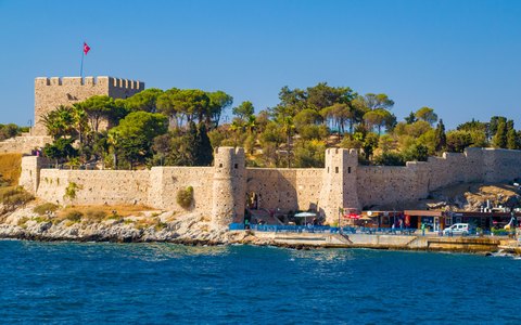 The stone fortress on Pigeon Island at Kuşadası with a Turkish flag, pine trees and blue water under a blue sky