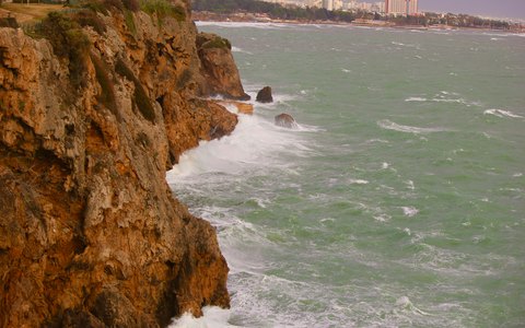 Golden rock cliffs above a rough green sea at Lara, with buildings along the coast in the distance