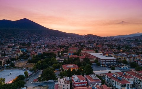 Aerial view of the town of Milas below dark mountains under a pink and orange sunset sky