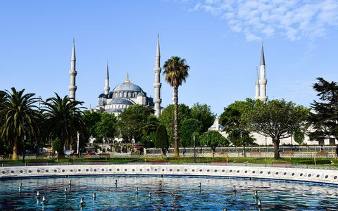 The domes and minarets of the Blue Mosque behind gardens, palm trees and a fountain, under a blue sky