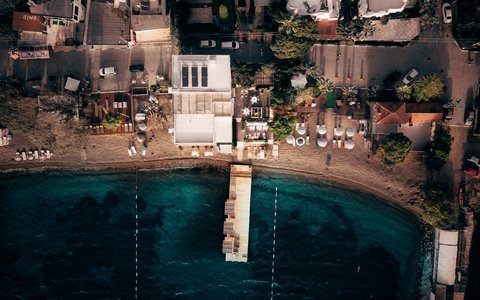 Overhead drone view of a small bay in Yalıkavak, with a jetty, a sandy beach with sunshades and buildings along the shore