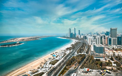 Aerial view of the Abu Dhabi Corniche with its beach, turquoise water and city towers, under a blue sky with streaks of cloud
