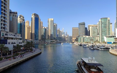 Skyscrapers lining the water of Dubai Marina with yachts and a promenade, under a clear blue sky
