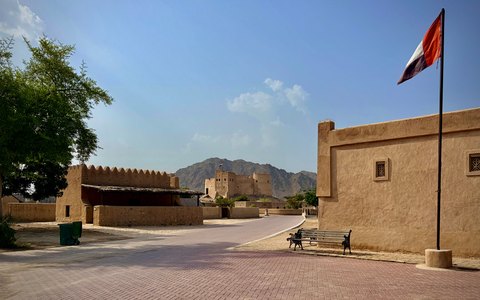 The mud-brick walls of Fujairah Fort with the UAE flag and mountains behind, in warm daylight
