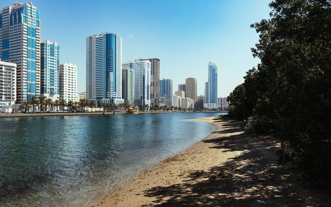 High-rise towers of Sharjah along the Al Majaz waterfront, with a small sandy shore and trees, under a clear sky