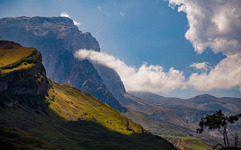 Ein sonnenbeschienener grüner Bergrücken vor hohen grauen Felswänden, mit einem weißen Wolkenband und strahlend blauem Himmel