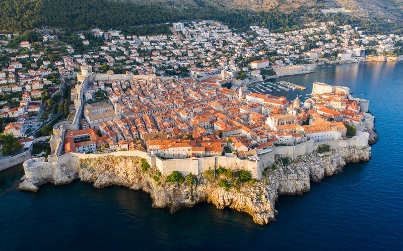 Luftaufnahme der ummauerten Altstadt Dubrovniks auf ihrer felsigen Landzunge, mit roten Dächern, dem Hafen und tiefblauem Meer