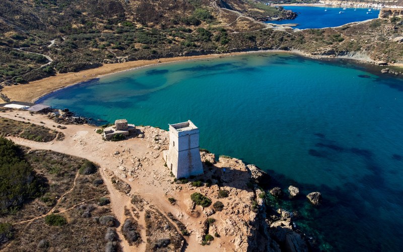 Luftaufnahme einer geschwungenen Sandbucht mit türkisfarbenem Wasser, einem quadratischen steinernen Wachturm auf der Landzunge und einer weiteren Bucht in der Ferne