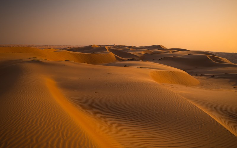 Gewellte goldene Sanddünen erstrecken sich unter einem warmen orangefarbenen Abendhimmel in die Ferne