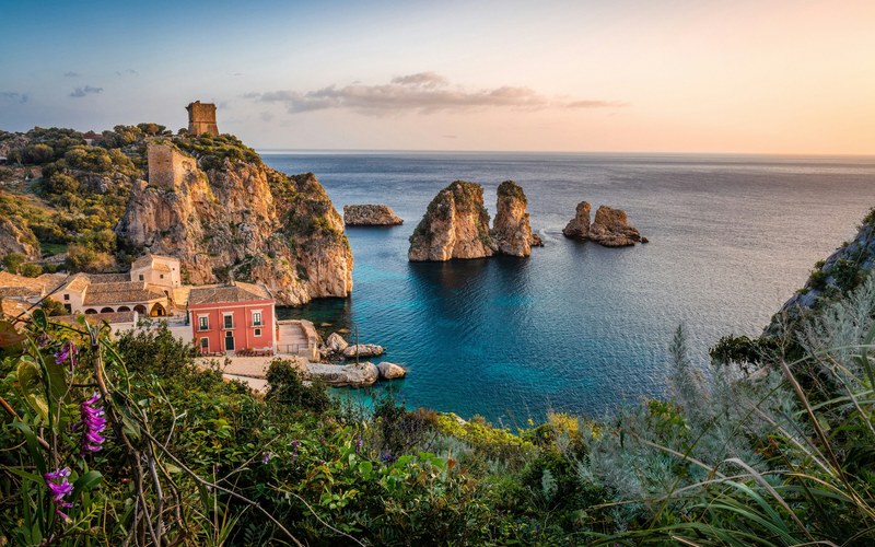 Eine alte Thunfischfanganlage mit einem Steinturm auf einer felsigen Landspitze, Felsnadeln im ruhigen blauen Meer und Wildblumen im Vordergrund im sanften Abendlicht